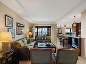 A modern living room with a sofa, chairs, a coffee table, and a TV. There is a kitchen area with a bar and a view of the ocean. - Private Lanai Ocean View Retreat | 3BR Sleeps 6 (Lahaina)