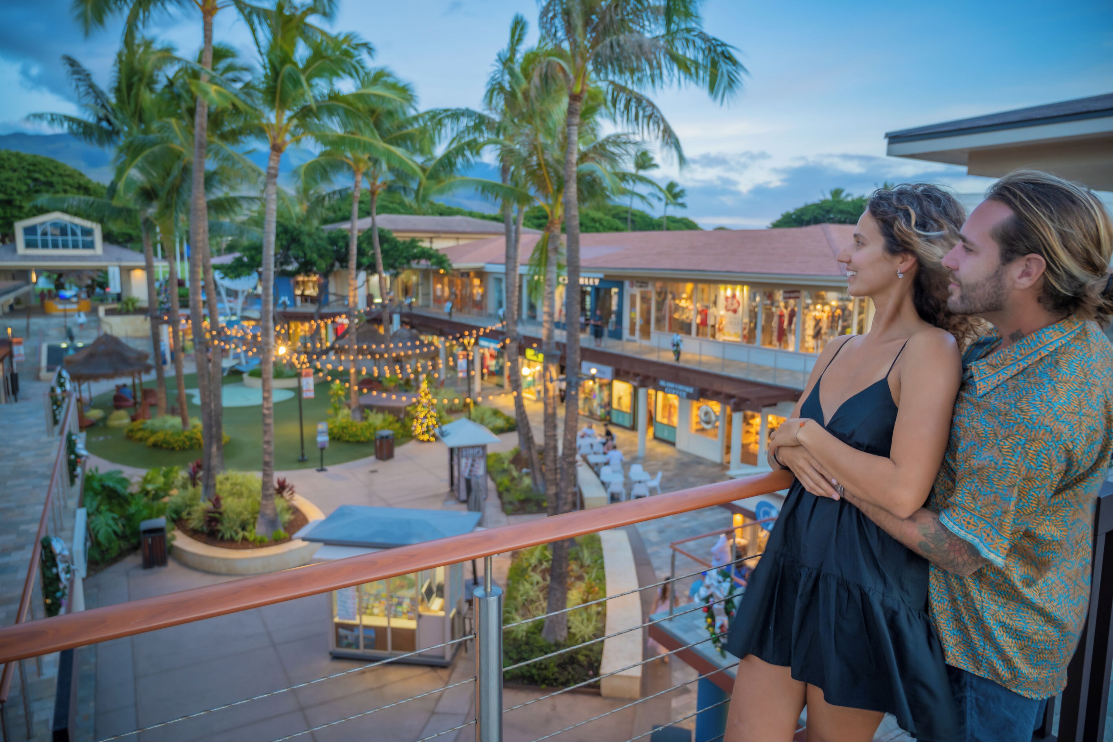 A couple standing on a balcony overlooking a resort area with palm trees and a lit-up dining area.