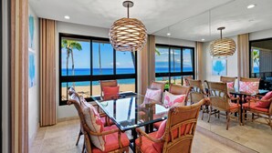 A dining area with a glass table, wicker chairs, and a view of the beach. - Wraparound Ocean Views | Sleeps 6 w/ Resort Amenities (Lahaina)