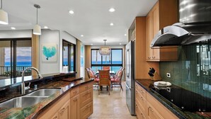 A modern kitchen with granite countertops, wooden cabinets, and a view of the ocean. - Wraparound Ocean Views | Sleeps 6 w/ Resort Amenities (Lahaina)