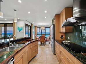 A modern kitchen with granite countertops, wooden cabinets, and a view of the ocean. - Wraparound Ocean Views | Sleeps 6 w/ Resort Amenities (Lahaina)