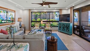 A spacious living room with a large window offering a view of a residential area and the ocean. There is a ceiling fan, a TV, and a seating area with a sofa and chairs. - Wraparound Ocean Views | Sleeps 6 w/ Resort Amenities (Lahaina)