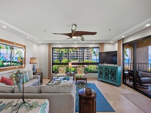 A spacious living room with a large window offering a view of a residential area and the ocean. There is a ceiling fan, a TV, and a seating area with a sofa and chairs. - Wraparound Ocean Views | Sleeps 6 w/ Resort Amenities (Lahaina)