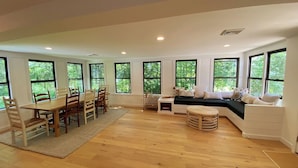 A spacious room with a dining table and chairs, a built-in bench with cushions, and large windows offering a view of greenery. - Renovated 3BR Home near  High Falls / Stone Ridge (Accord)