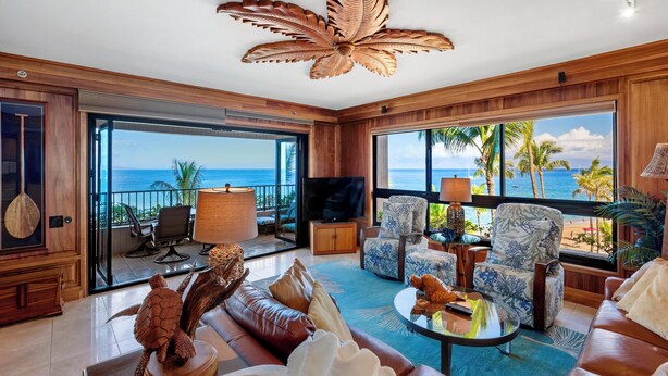 Ein geräumiges Wohnzimmer mit großem Fenster und Meer- und Palmenblick. Es gibt ein Ledersofa, einen Couchtisch und einen Fernseher.