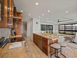 A modern kitchen with wooden cabinets, a marble countertop, and a large window offering a view of the city. - Front-Row to the Pacific | Oceanfront Living at Kaanapali Alii (Lahaina)