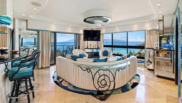 Un elegante salón con una mesa de centro de vidrio curvo, un sofá con cojines azules y vistas al mar.