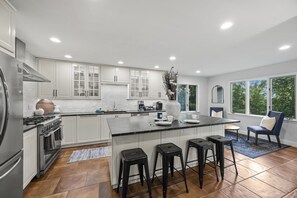 A modern kitchen with white cabinets, a central island with stools, a large window, and a view of greenery. - Asheville Mtn Chalet: Spa & Theater (Asheville)