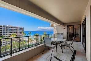 Ein Balkon mit Tisch und Stühlen, der auf einen Strand und ein Meer mit Gebäuden im Hintergrund blickt.