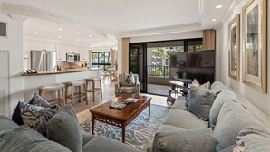 Un salon moderne avec un grand canapé, une table basse et un téléviseur. Il y a un coin cuisine avec des meubles blancs et un réfrigérateur. La pièce est dotée d’une grande fenêtre donnant sur des palmiers.
