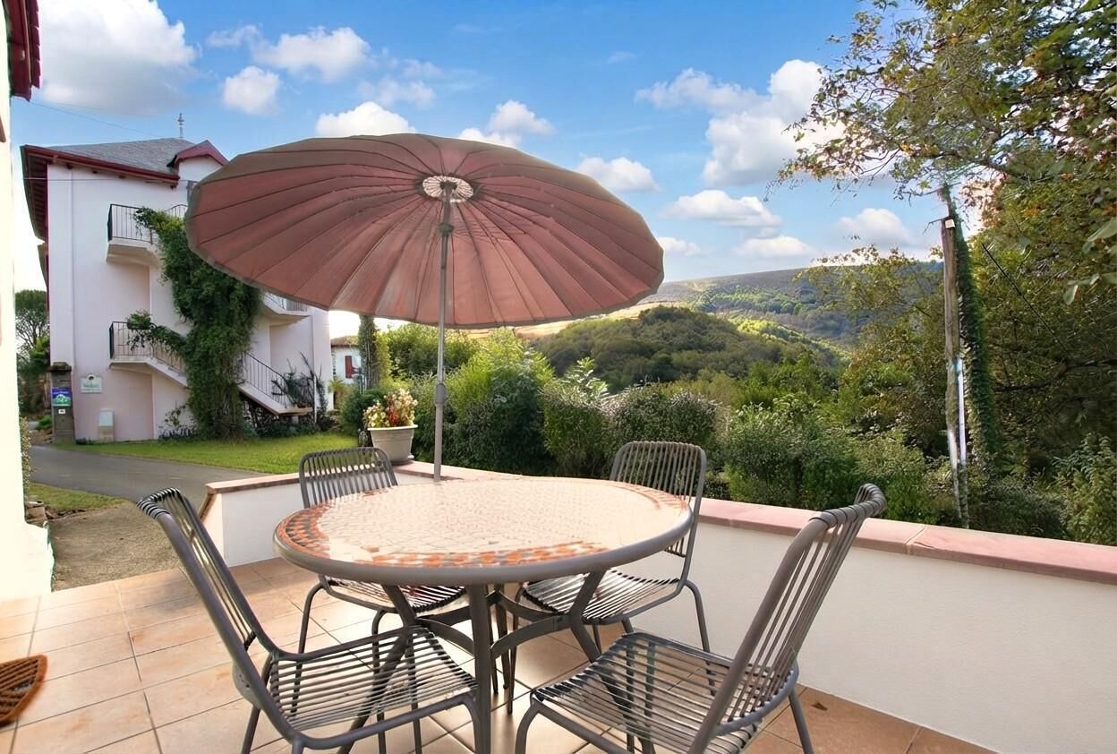 An outdoor patio with a round table and chairs, an umbrella, and a view of a hillside.