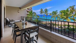 Een balkon met een tafel en stoelen, uitzicht op een strand met palmbomen en een gebouw op de achtergrond.