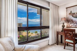 A room with a large window overlooking a beach and ocean, a wooden chair, a lamp, and a framed artwork on the wall. - Stunning Ocean View - Professionally Managed (Lahaina)