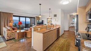 Une cuisine moderne dotée d’un îlot central, un coin repas avec un canapé et des chaises, et un salon avec une télévision et une vue sur l’extérieur.