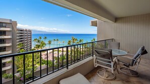 Un balcon avec vue sur l’océan et les palmiers, une table avec des chaises et un petit coin salon.
