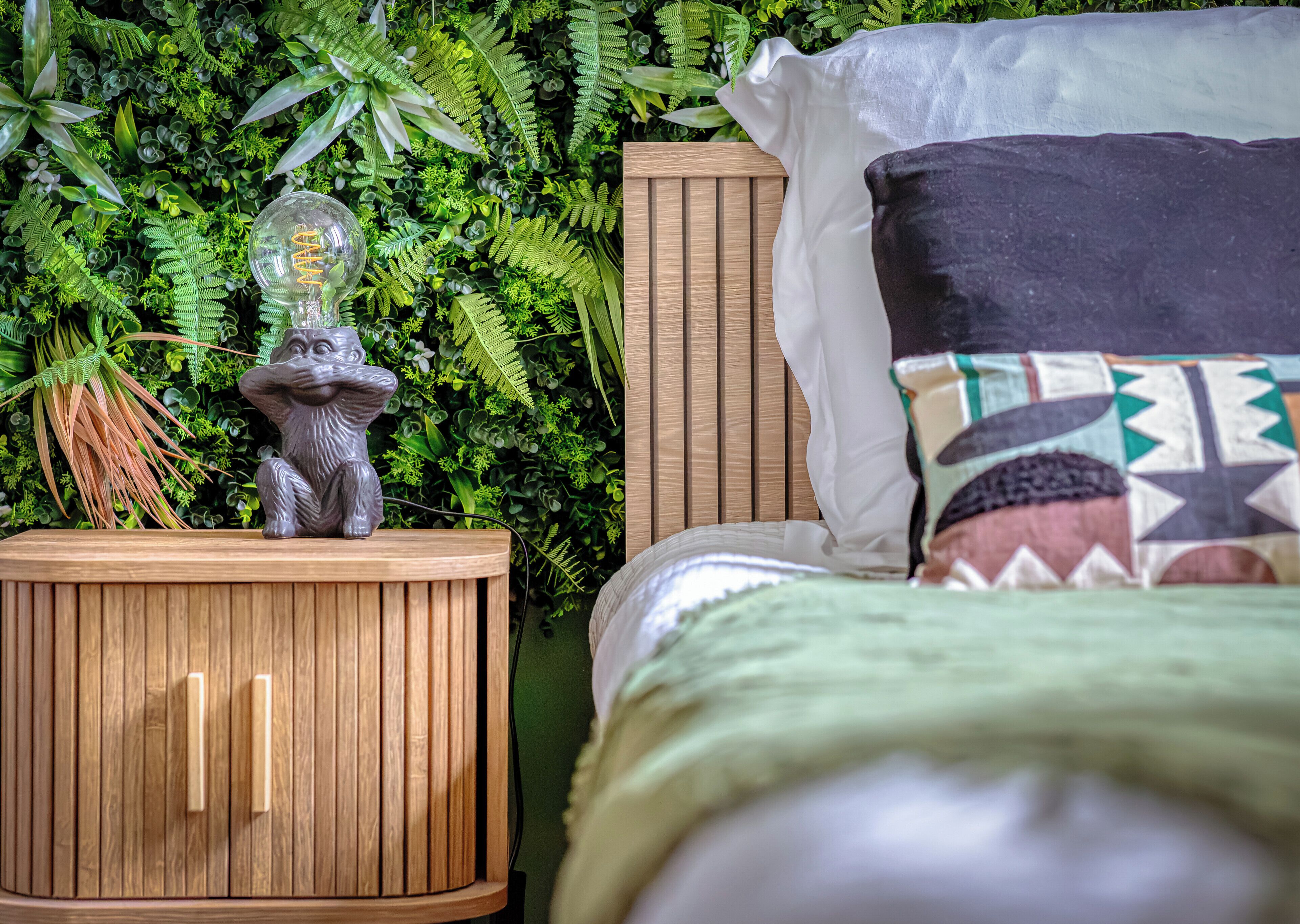 A bed with a patterned pillow, a nightstand with a decorative lamp, and a lush green plant wall.