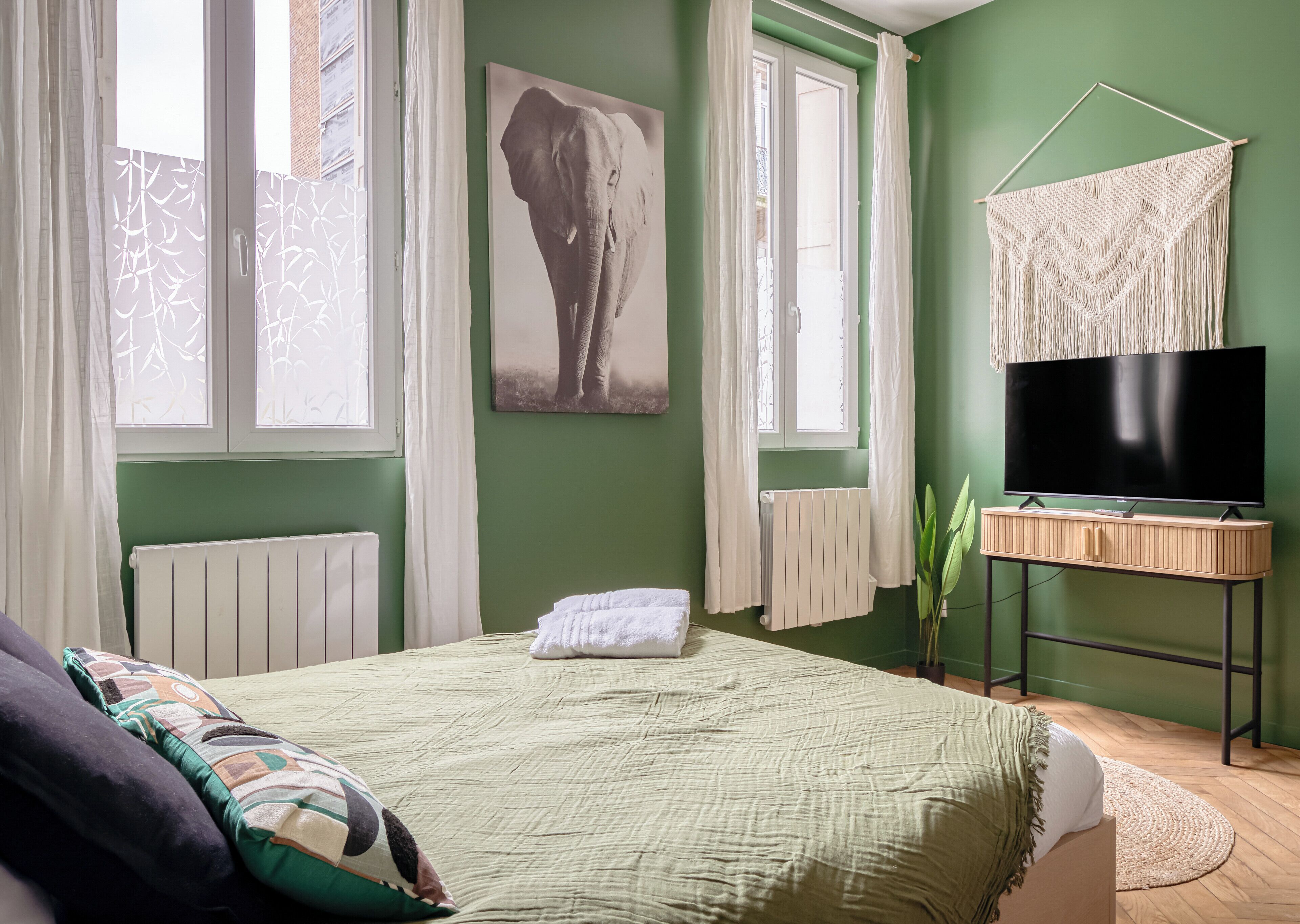 A bedroom with a bed, a television on a stand, a radiator, and a window with curtains.