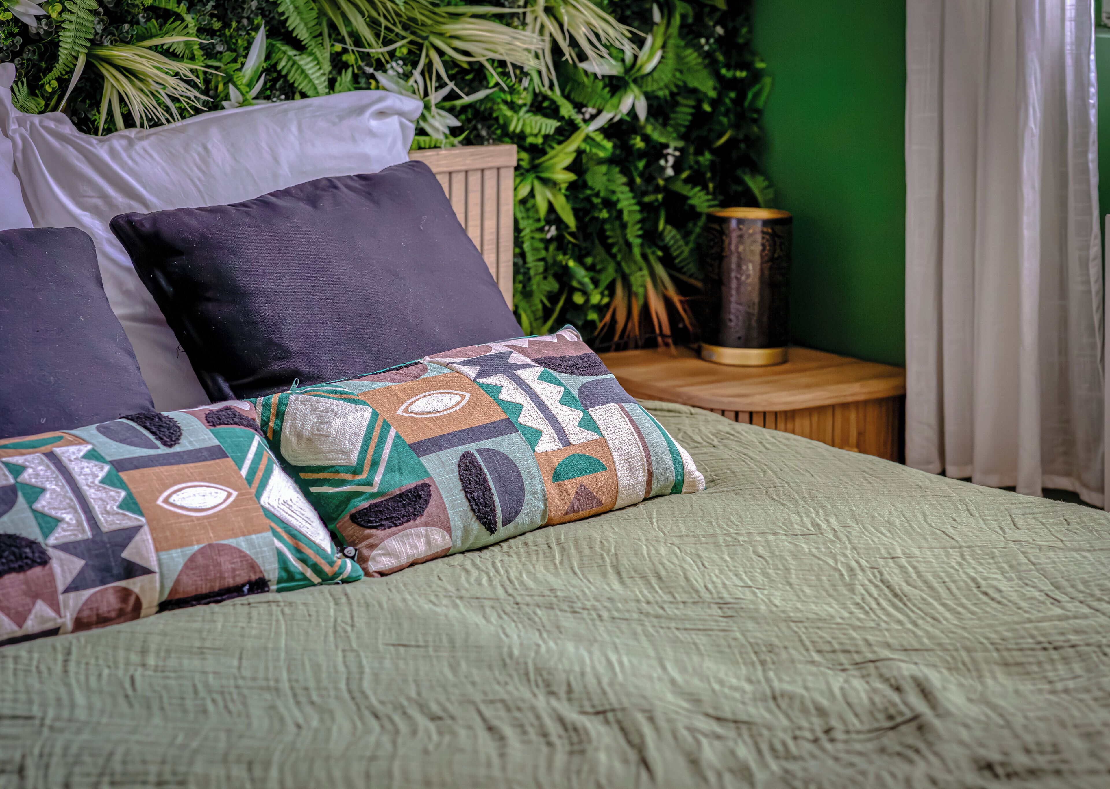 Un letto con cuscini decorati, un comodino in legno e una parete verde con piante.