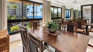Un comedor amplio con una gran mesa de madera y sillas, ventanas grandes con vistas a palmeras y un edificio, y ventiladores de techo.
