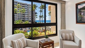 Habitación de hotel con dos sillones, una pequeña mesa de madera y un ventanal que ofrece vistas de un edificio hotelero, palmeras y el océano.