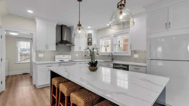 Une cuisine moderne avec des meubles blancs, un grand îlot central doté de tabourets, un éclairage suspendu et une vue sur l’extérieur.