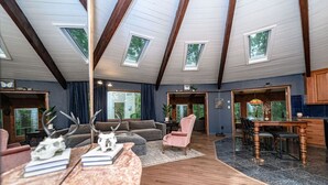 A spacious living area with a high ceiling, wooden beams, and skylights. There is a cozy seating area with a sofa and armchairs, a dining table, and a kitchen area visible through an open doorway.