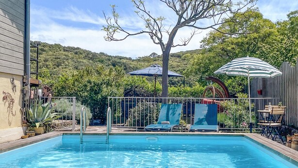 A swimming pool with lounge chairs and umbrellas, surrounded by a fence and greenery.