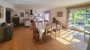 A spacious kitchen with wooden flooring, a dining area with a table and chairs, and a large window with a view of greenery.