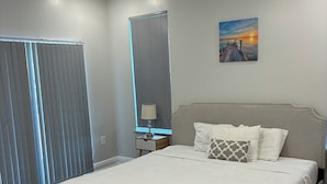 A bedroom with a large bed, a nightstand, a lamp, a ceiling fan, and a window with blinds.