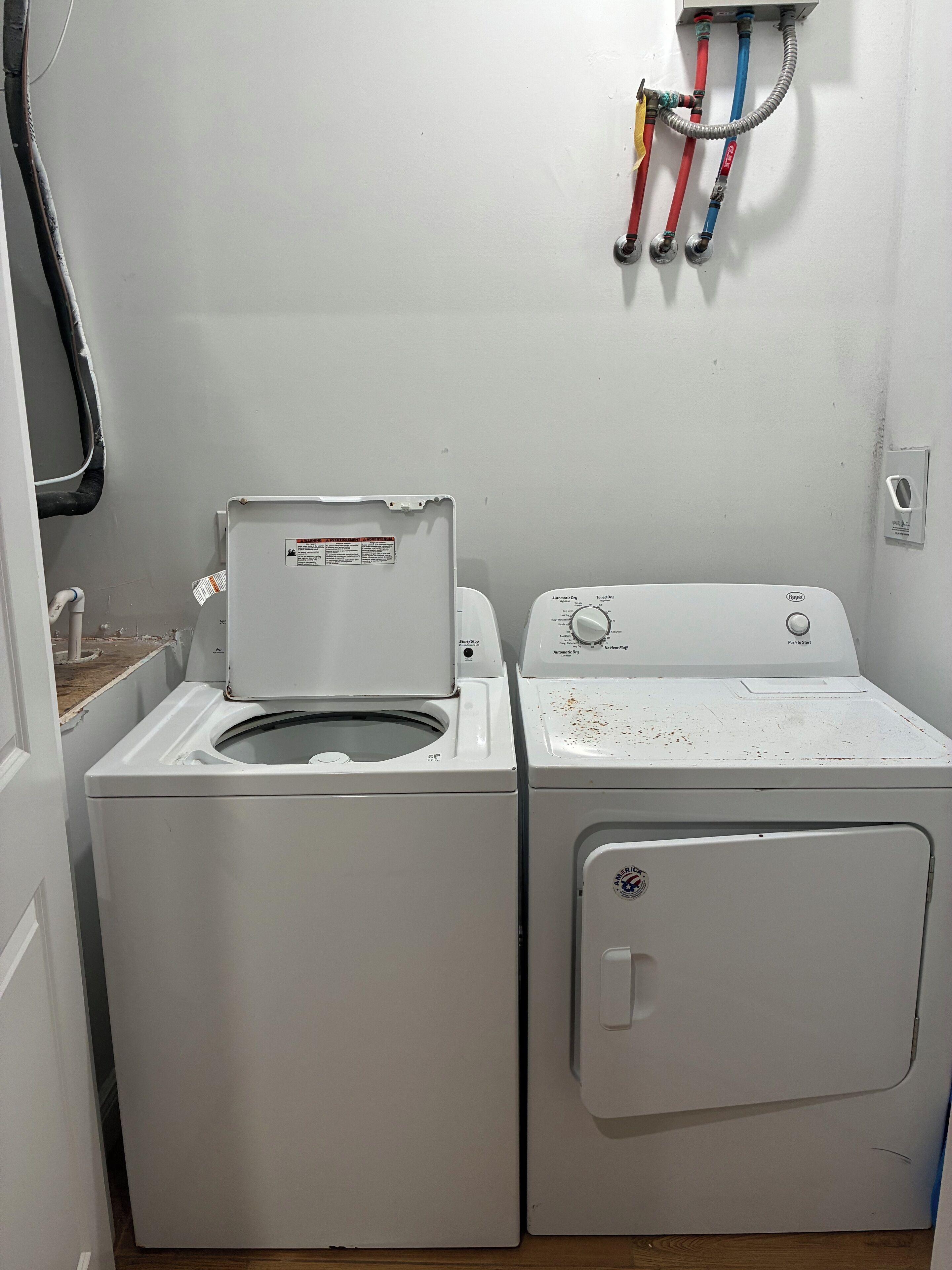 Une buanderie équipée de deux machines à laver blanches, d’une prise électrique murale et d’une installation de plomberie visible.