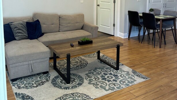 A living room with a sofa, coffee table, patterned rug, wooden flooring, and a ceiling fan.