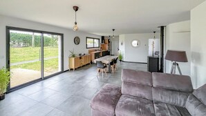 Una sala de estar moderna con un sofá gris, una zona de comedor con una mesa y sillas de madera, y una cocina con muebles de madera y un ventanal grande.