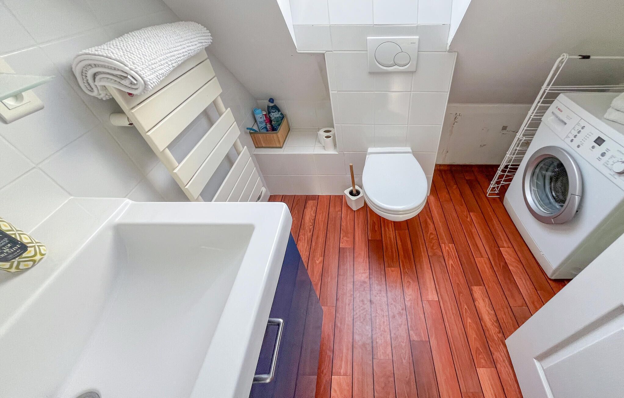 A compact bathroom with a wooden floor, a white toilet, a washing machine, and a bathtub.