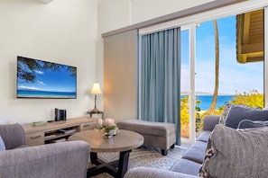 Ein Hotelzimmer mit einer Couch, einem kleinen Tisch mit Lampe, einem Fernseher an der Wand und Meerblick durch das Fenster.
