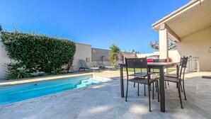 Un área de piscina con mesa y sillas, rodeada de follaje y un cielo azul despejado.
