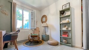 Una habitación con una silla de madera, una pequeña mesa redonda, una lámpara, una estantería y una ventana con cortinas.