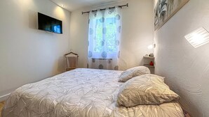 Un dormitorio con cama, televisor montado en la pared, ventana con cortinas y una mesita de noche con lámpara.