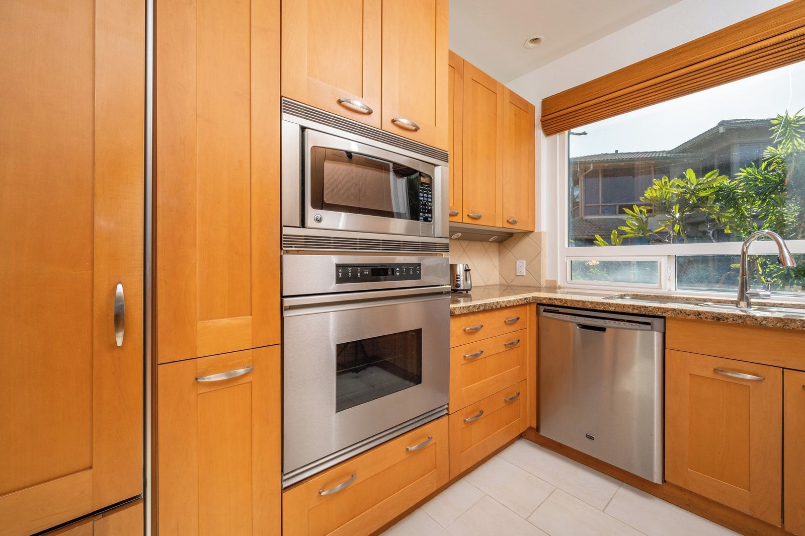A kitchen with wooden cabinets, stainless steel appliances, a window with a view of greenery, and a tiled floor.