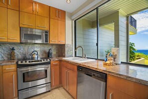 A modern kitchen with stainless steel appliances, wooden cabinets, a granite countertop, and a view of the ocean. - Kapalua Bay | Ocean View 6 BR, Sleeps 20 | Car Incl. w/ 6+ Nights | KBV ML-2059 by KBM (Lahaina)