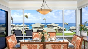 Comedor con una mesa dispuesta para cuatro personas, ventanas amplias con vistas a la costa, casas y palmeras.