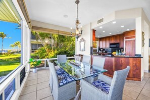 A modern kitchen with wooden cabinets, a glass island, and a dining area with wicker chairs and a glass table. - Kapalua Bay | Ocean View 4 BR, Sleeps 14 | Car Incl. w/ 6+ Nights | KBV ML-2068 by KBM (Lahaina)