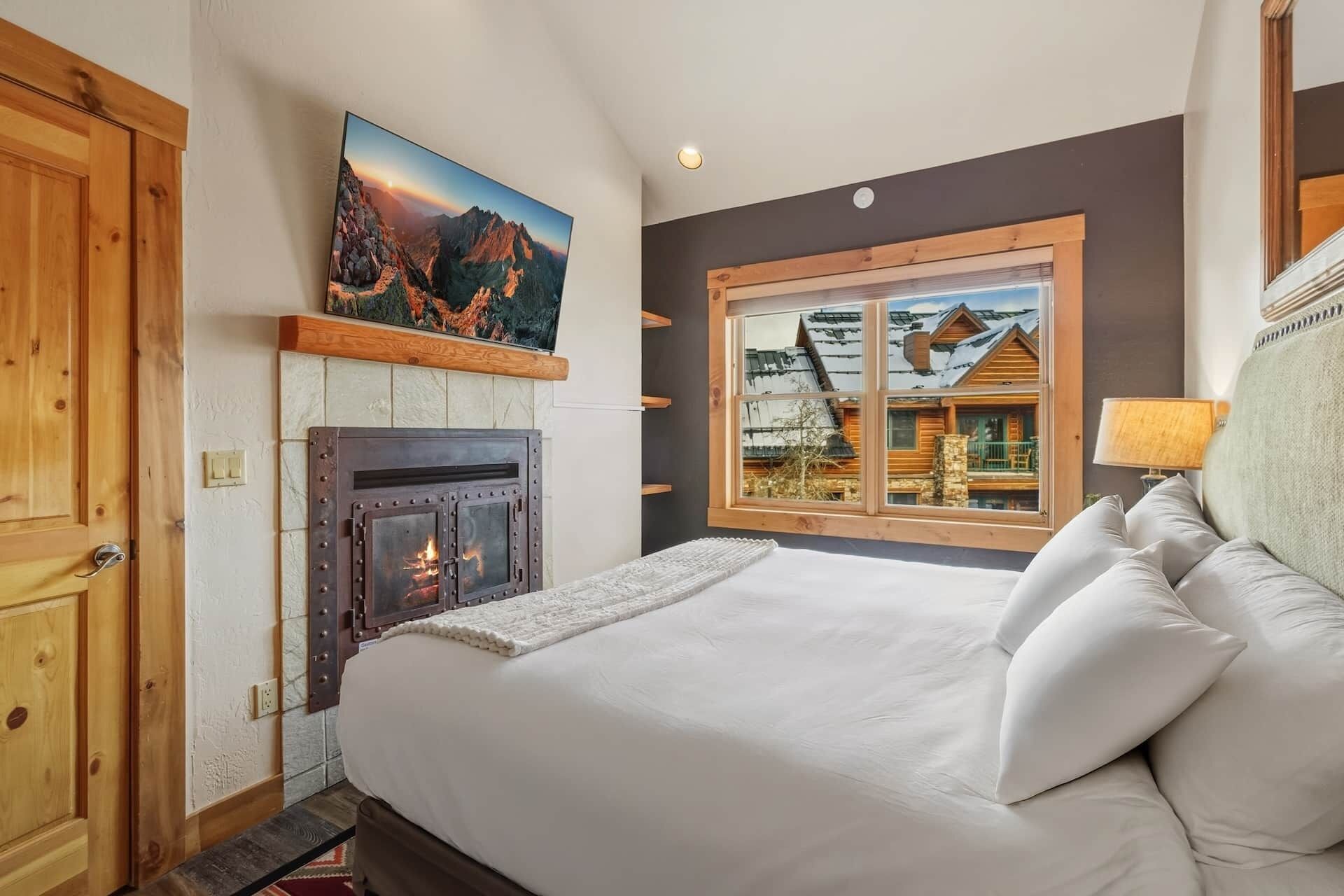 Ein Schlafzimmer mit Kamin, einem großen Fenster mit Blick auf eine Berghütte und einem Bett mit Kissen.