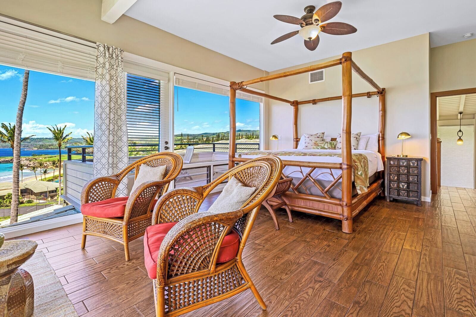 A four-poster bed with a view of the ocean, wicker chairs with cushions, and a ceiling fan.
