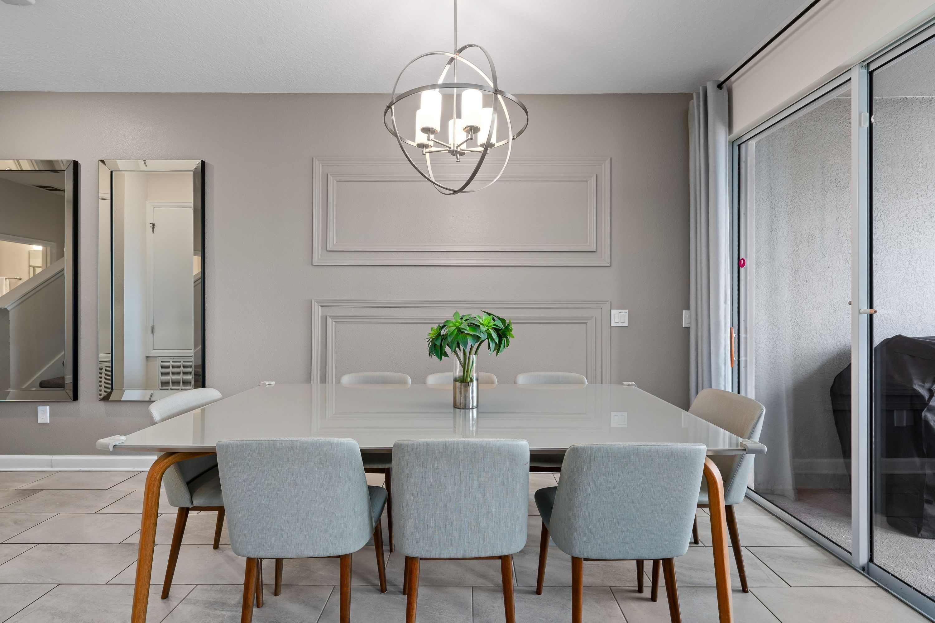 A modern dining room with a large table, four chairs, a chandelier, and a plant.