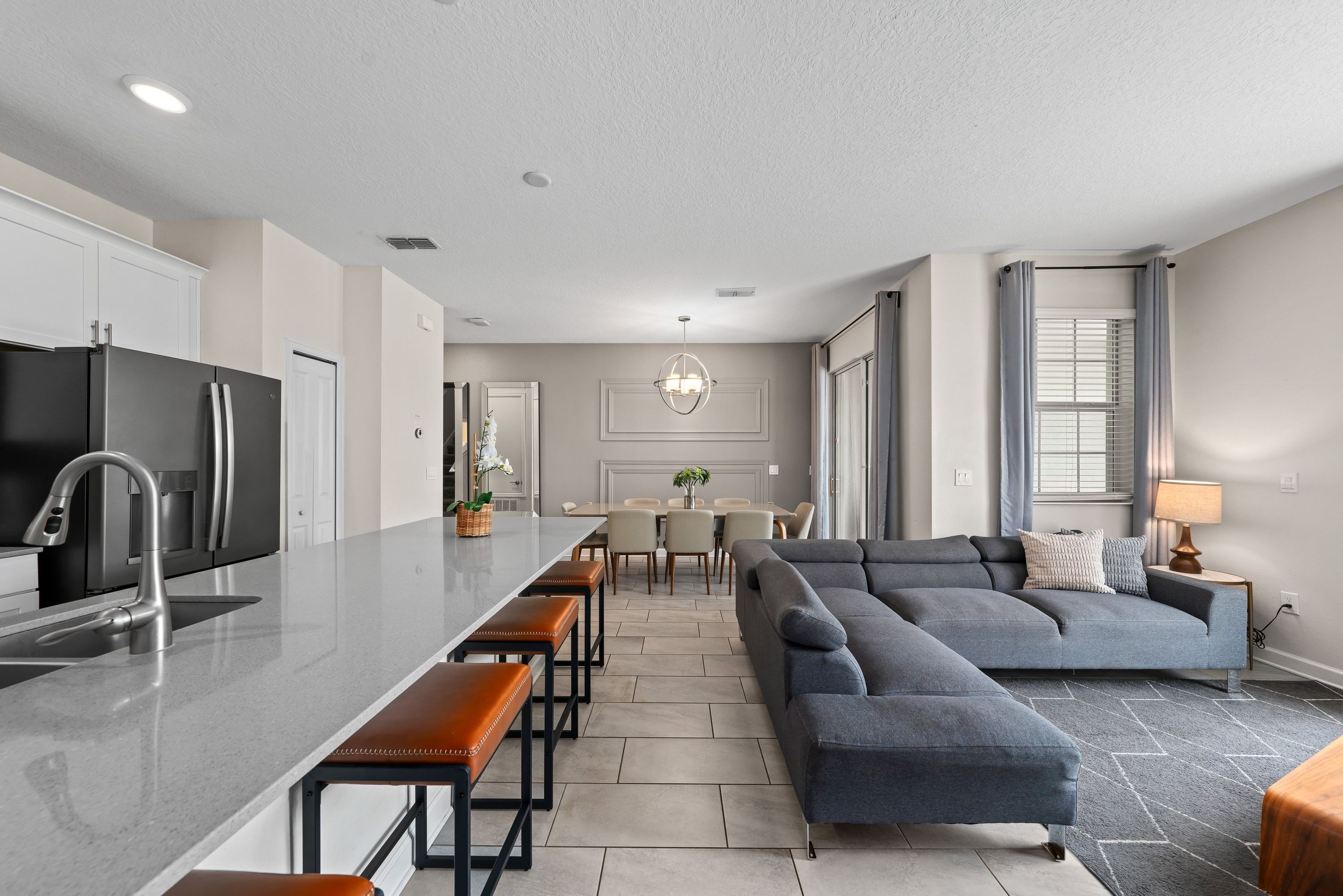 A modern kitchen with a central island, bar stools, a dining area with a table and chairs, and a living area with a sofa and a lamp.