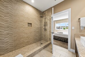A modern bathroom with a herringbone-patterned wall, a glass-enclosed shower, and a view of a bedroom with a bed and window. - Bright and Modern Townhome at Magic Village - Superb Disney Location (Kissimmee)