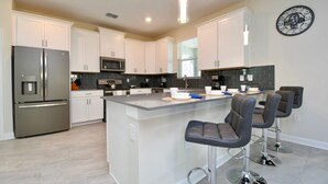 A modern kitchen with white cabinets, stainless steel appliances, a central island with bar stools, and a clock on the wall. - Modern Oasis Near Shopping n Golf Course w Pool (Davenport)