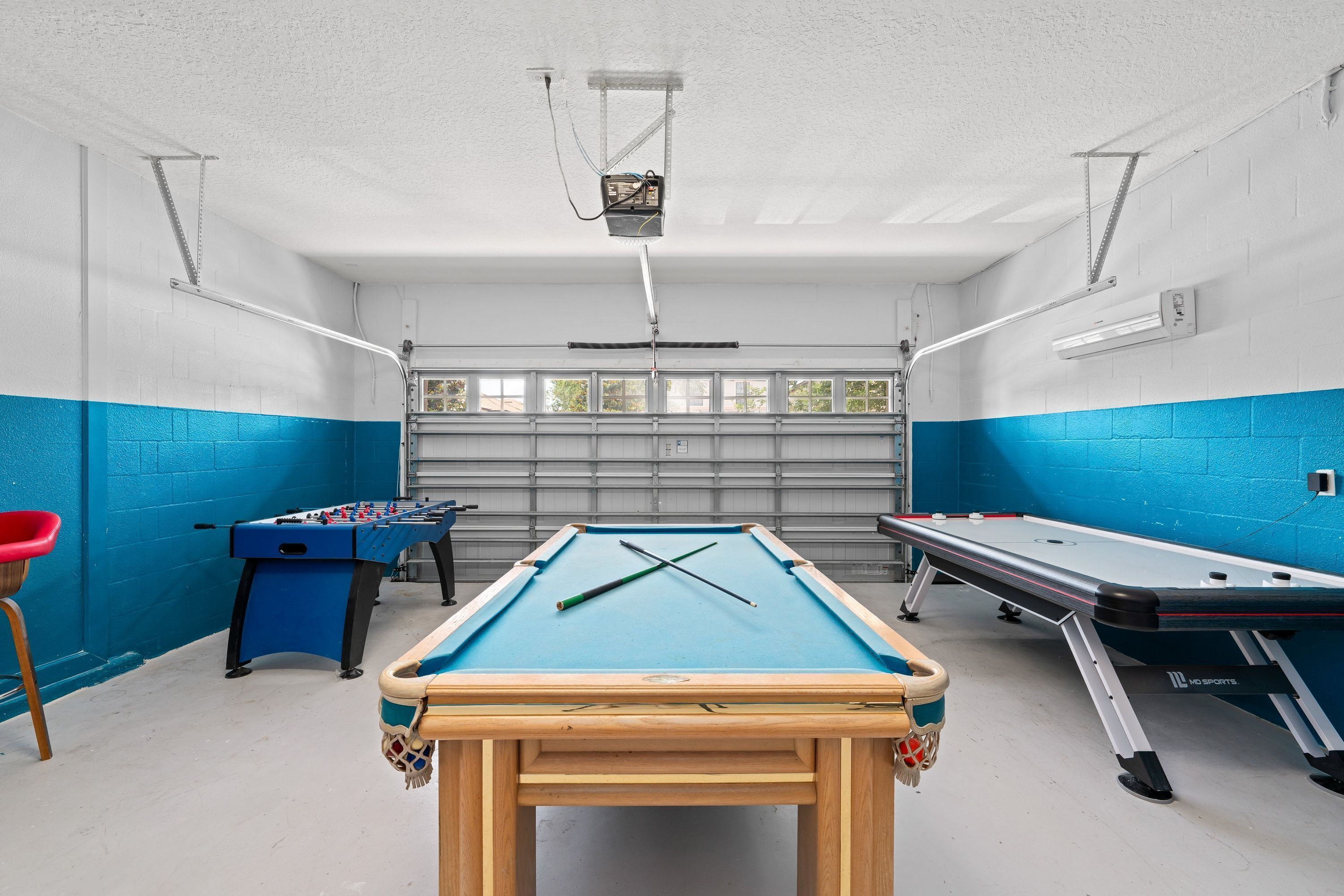Een pooltafel met een blauwe viltkleed, een tafelvoetbaltafel en een biljarttafel in een ruimte met blauwe muren en een garage deur op de achtergrond.