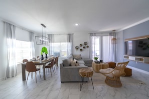 A modern living room with a grey sofa, dining table, and chairs. There is a TV mounted on the wall and a hanging light fixture. - Champions Gate Retreat Private Pool Top Resort (Davenport)
