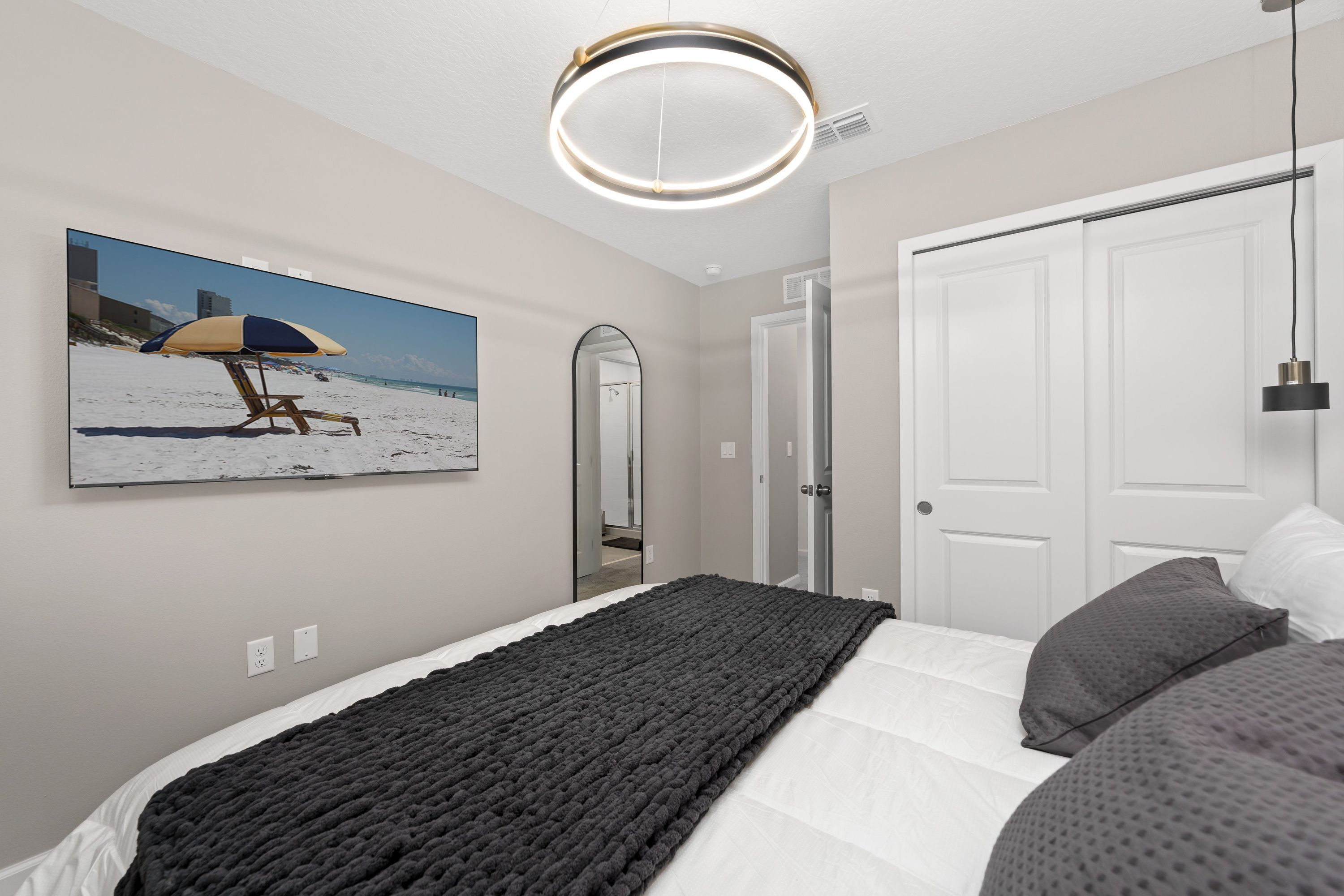 A bedroom with a bed, a dark blanket, a beach-themed painting, a mirror, and a door.